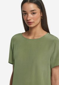 Olivgrön kortärmad blus med rund halsringning. Slät, något glänsande tyg. Enkelt design utan synliga mönster eller accenter.