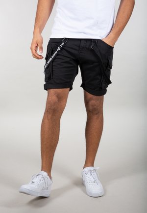 Man draagt zwarte cargoshorts met riemen en witte sneakers, staat met één hand in de zak tegen een effen achtergrond.