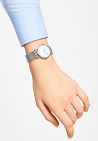 Zilveren gaasbandhorloge met een witte wijzerplaat, minimalistische zilveren uurmarkeringen, liggend op een hand met een blauwe shirtboord.