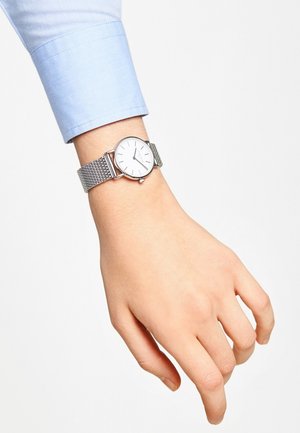 Montre avec un bracelet en maille argentée, un cadran blanc et des index d'heure argentés minimalistes, posée sur une main avec un poignet de chemise bleue.