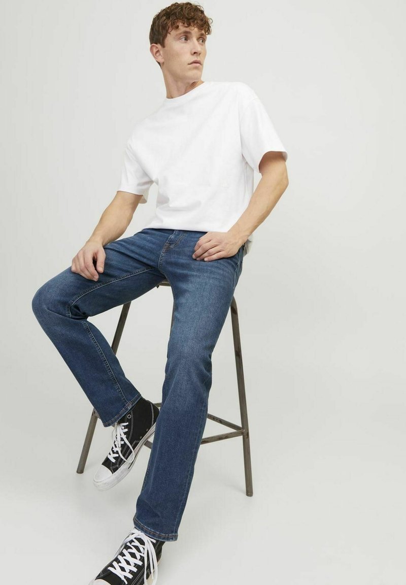 T-shirt blanche à manches courtes associée à un jean bleu foncé et des baskets noires, assis sur un tabouret en métal contre un fond clair.