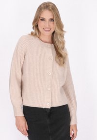 Beige gebreide cardi met een geribbelde textuur, ronde hals en sluiting met vijf knopen. Lange mouwen lopen smal toe bij de manchetten. Gecombineerd met een donkere rok.