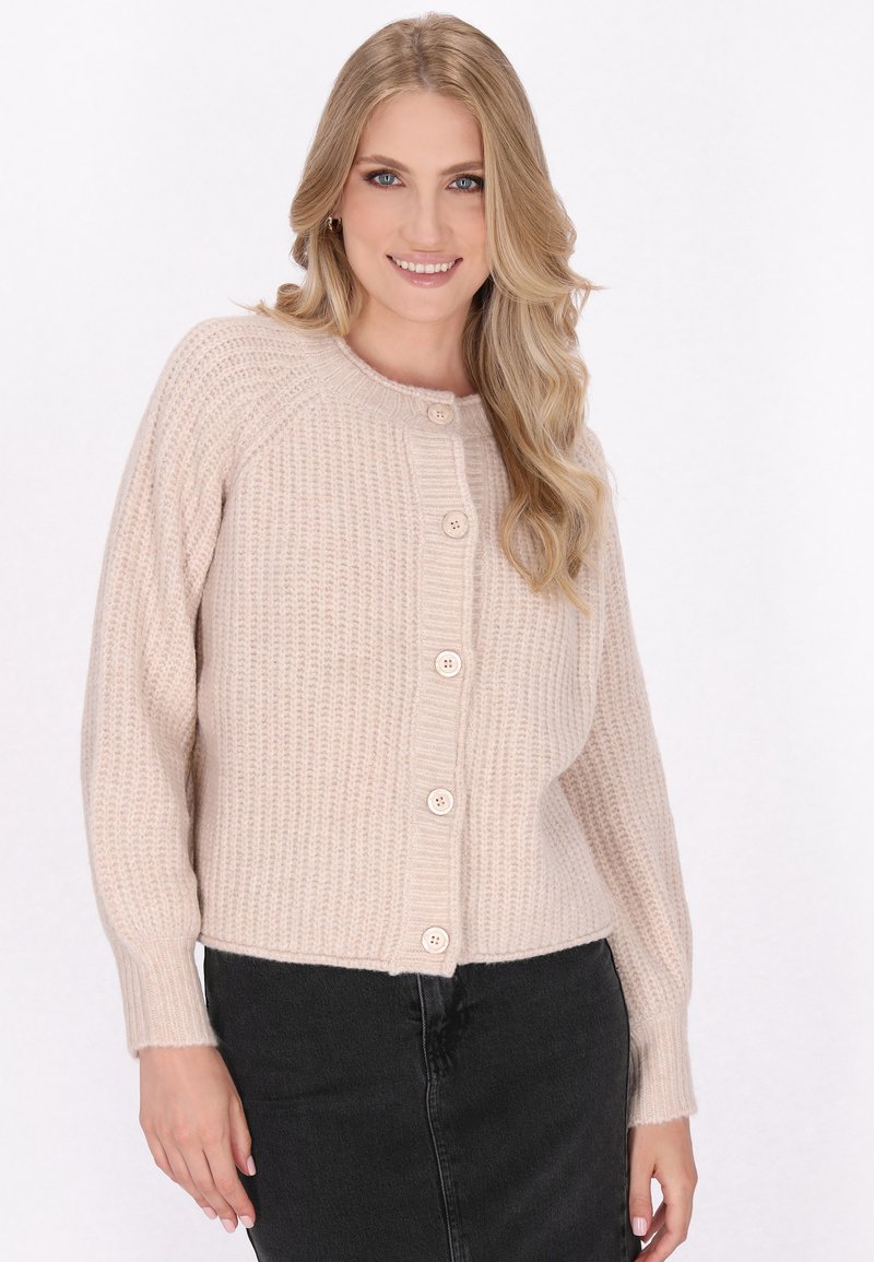 Beige gebreide cardi met een geribbelde textuur, ronde hals en sluiting met vijf knopen. Lange mouwen lopen smal toe bij de manchetten. Gecombineerd met een donkere rok.
