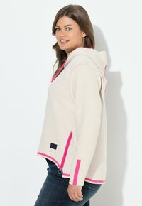 Sweatshirt beige à capuche avec des accents roses, doté d'une poche frontale et d'une fermeture éclair sur le côté. Texture douce et coupe décontractée, adapté pour un usage quotidien.