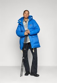 Vrouw in een blauwe gewatteerde jas en zwarte broek, die zwarte wandelstokken vasthoudt, staand tegen een effen lichte achtergrond.