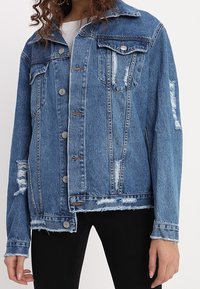 Sininen farkkutakki, jossa on kuluneita yksityiskohtia, napitus, kaksi rintataskua ja klassinen kaulus. Takki on rento malli ja siinä on frayed-helma.