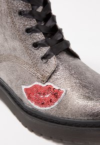 Botte argentée métallique à lacets noirs avec un patch lèvres rouges à sequins sur le côté extérieur au-dessus de la semelle noire.