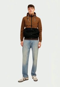 Brown and black zip-up hoodie with a drawstring hood, paired with light blue straight-leg jeans and beige sneakers. Smooth texture.