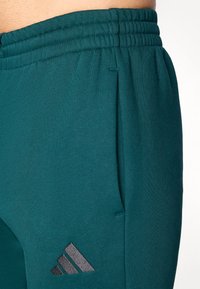 Turkizne atletske hlače z elastičnim pasom, ki imajo diskreten stranski žep in sivi Adidasov logotip na levi nogi. Gladka tekstura.