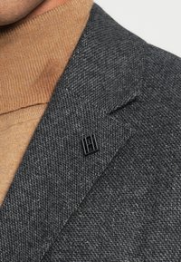 Colarinho de blazer texturizado cinzento com um alfinete metálico preto. Por baixo, um suéter bege às riscas visível no pescoço.