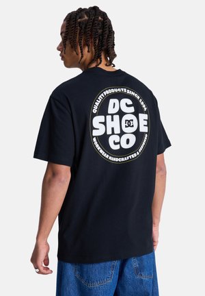 Zwart katoenen T-shirt met een groot rond wit logo op de rug met tekst, korte mouwen en een relaxte pasvorm. Denim shorts zichtbaar.