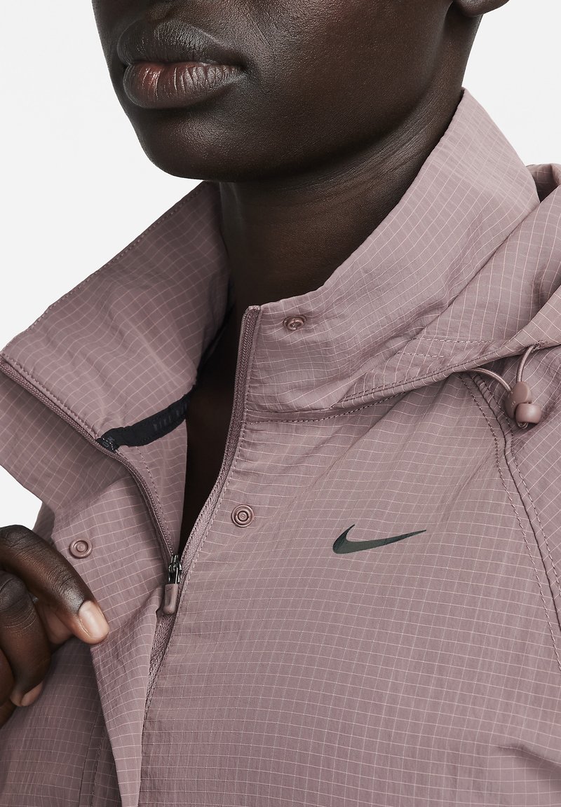 Nike-jack in licht mauve, met een ruitpatroon, een voorzak met ritssluiting, hoge kraag en verstelbare koordjes. Inclusief zwart logo-accent.