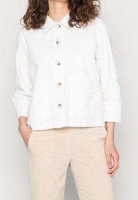 Chaqueta blanca de botones con cuello, dos bolsillos frontales, mangas de tres cuartos y una tela texturizada. Combinada con pantalones beige.