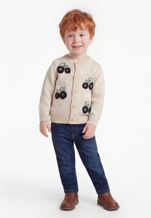Jeune garçon roux souriant, portant un cardigan beige avec des motifs de tracteur, un jean bleu et des bottes marron, debout sur fond blanc.