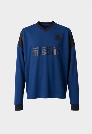 Homme portant une chemise bleue à manches longues avec des accents noirs et un texte, associée à un jean large bleu et des chaussures foncées, debout sur un fond blanc.