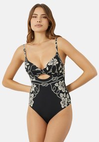 Vrouw in een zwarte badpak met crèmekleurige bloemen- en touwmotieven, dunne bandjes en een kleine uitsnijding onder de buste.
