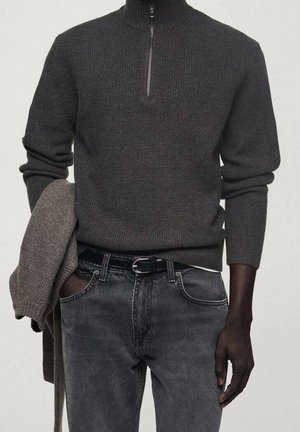 Pull en maille gris foncé avec un col montant et une fermeture éclair à mi-hauteur, associé à un jean noir décoloré et à un cardigan gris clair drapé sur le bras.