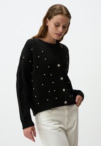 Zwarte gebreide cardigan met een V-hals, textuurpatronen, parelachtige versieringen en een knoopsluiting, gecombineerd met witte broeken.