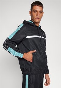 Chaqueta deportiva negra con detalles en turquesa y blanco, que cuenta con capucha, bolsillos laterales, puños elásticos y un diseño ajustado.