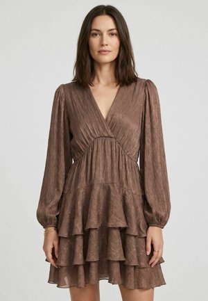 Femme portant une robe marron à manches longues, à volants, avec un col en V et un tissu texturé, debout devant un fond clair uni.