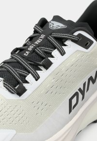 Ελαφρύ παπούτσι τρεξίματος σε λευκό και μαύρο, με υφή στην επάνω πλευρά, αντίθετα κορδόνια και εμφανές λογότυπο 'DYN' στο πλάι.