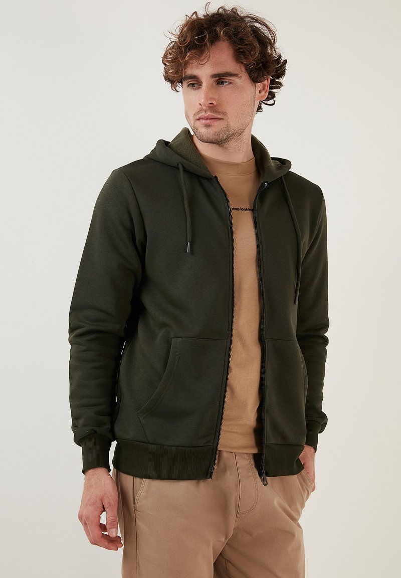 Maslinasta zip-up dukserica izrađena od mekanog materijala, s kapuljačom na šnuru, prednjim džepovima i rebrastim manžetama. Nosi se preko bež majice.