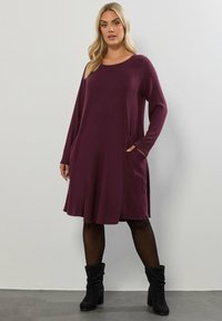 Burgundowa sukienka z długim rękawem, okrągłym dekoltem, bocznymi kieszeniami i rozkloszowanym dołem. Tkanina wydaje się miękka o lekko fakturowanym wykończeniu.