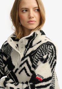 Young woman in a black and white patterned fleece jacket with snap buttons and "Superdry" logo on sleeve, looking sideways against white background.