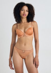 BH- en stringset in perzik met kantdetails. BH heeft gladde cups, verstelbare bandjes en een sluiting aan de voorkant. De string heeft een kantafwerking met een klein accent.