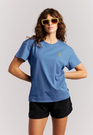 Vrouw met krullend haar die gele zonnebril, een blauw t-shirt met kleine gouden tekst en zwarte short draagt, staat met handen in de zij.