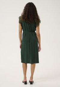 Zelené šaty s krátkými krajkovými rukávy, staženým pasem a délkou pod kolena. Hladká textura s jemně splývavou siluetou, nošené s sandály na podpatku.