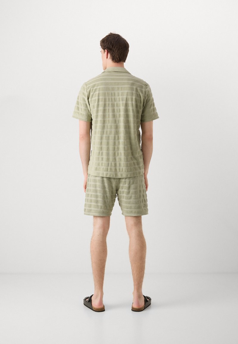 Hellgrünes, strukturiertes kurzärmliges Hemd und passende Shorts mit horizontalen Streifen. Lässiges Design, kombiniert mit dunklen Sandalen.