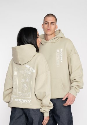 Hanorac oversized bej, cu un buzunar frontal, având desene grafice albe și text pe spate. Textură moale și design lejer.