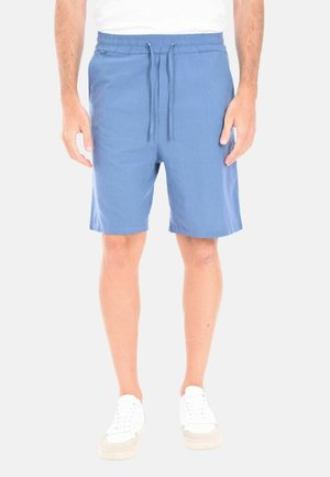 Uomo che indossa pantaloncini casual blu con coulisse e scarpe da ginnastica bianche, in piedi davanti a uno sfondo semplice.