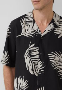 Zwart shirt met korte mouwen, versierd met een natuur geïnspireerd beige bladerenpatroon, met een notch-kraag en voorknopen voor sluiting.