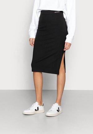 Kvinde iført en sort midi-nederdel med sideslids, hvid langærmet top og hvide sneakers med sorte detaljer.