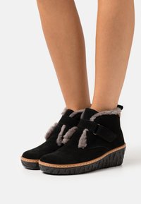 Black suede ankle boots with a wedge outsole, featuring gray fur lining, adjustable straps, and stitched detailing along the sole edge.