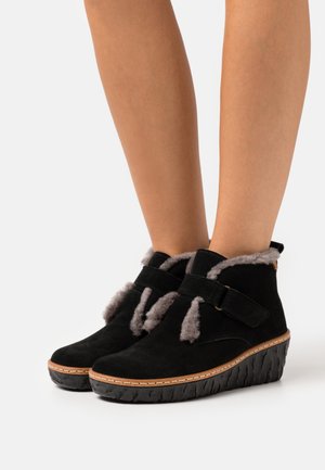 Stivaletti neri in suede con suola a zeppa, dotati di foderatura in pelliccia grigia, cinturini regolabili e dettagli cuciti lungo il bordo della suola.
