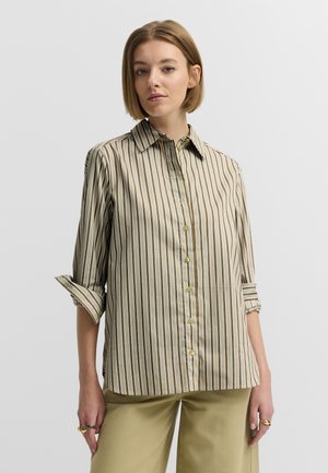Woman with short blonde hair wearing a beige and brown vertical striped button-up shirt with rolled sleeves and light khaki pants.