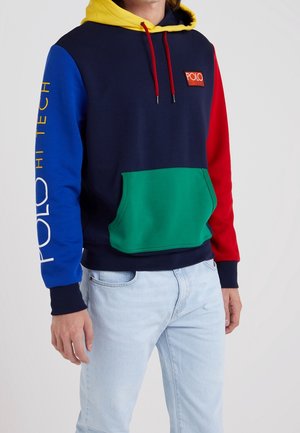 Mężczyzna w kolorowej bluzie z kapturem w żółtym kolorze, niebieskim lewym rękawem z napisem "Polo Hi Tech", czerwonym prawym rękawem, zieloną kieszenią z przodu oraz jasnymi dżinsami.