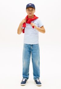 Αγόρι που φορά μπλε καπέλο, λευκό μπλουζάκι, κόκκινο πουλόβερ ριγμένο στους ώμους, μπλε τζιν και ναυτικό μπλε αθλητικά παπούτσια, στέκεται μπροστά σε λευκό φόντο.