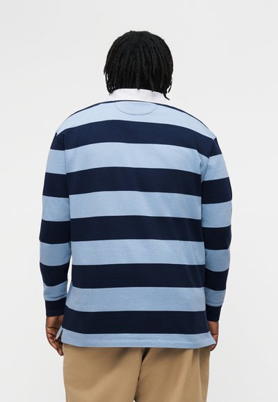 Személy sötét raszta frizurával, hosszú ujjú rugby ingben, tengerészkék és világoskék széles vízszintes csíkokkal, bézs nadrágban, hátulnézetben.