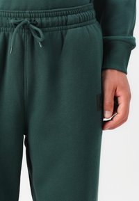 Mørkegrønne joggingbukser lavet af blød bomuldsblanding, med elastisk talje og snor, samt et lille brunt logo-patch på siden.