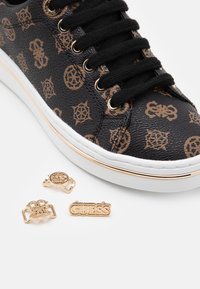 Schwarze synthetische Sneakers mit strukturiertem Muster und goldenen Akzenten. Enthält goldene Markenlogos und dekorative Pins neben dem Schuh.