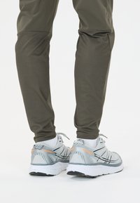 Persoon in olijfgroene broek en witte hardloopschoenen met oranje "Endurance" tekst, staande op een witte achtergrond.