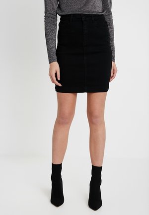 Vrouw draagt een zwarte denim minirok, gecombineerd met een sprankelende top met lange mouwen en zwarte enkellaarzen, staand tegen een effen achtergrond.