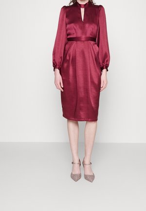 Woman wearing a knee-length burgundy satin dress with long puffed sleeves and a keyhole neckline, paired with glittery ankle-strap heels.