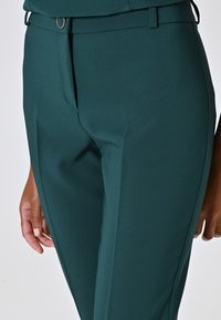 Türkisfarbene Damenhose aus einem glatten, dehnbaren Stoff, mit flachem Bund, Gürtelschlaufen und dezenten Falten entlang der Beine.