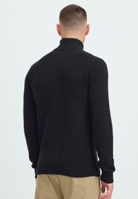 Mężczyzna z krótkimi włosami, ubrany w czarny sweter z golfem i beżowe spodnie, zwrócony plecami na tle białego, jednorodnego tła.