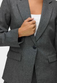 Pelēks teksturēts žakete ar vienu melnu pogu, iezīmētām atloklapām un divām priekšējām kabatām. Valkāta virs baltas topiņa.